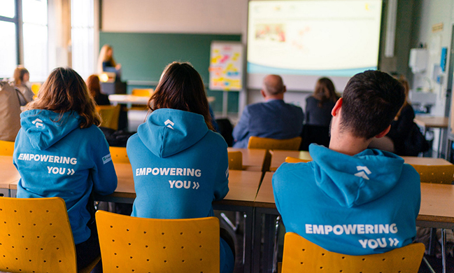 Drei Personen in blauen Kapuzenpullovern, auf denen "Empowering you" steht, sitzen in einem Seminarraum und hören einem Vortrag zu (© Ludmilla Parsyak Photography)  Drei Personen in blauen Kapuzenpullovern, auf denen "Empowering you" steht, sitzen in einem Seminarraum und hören einem Vortrag zu