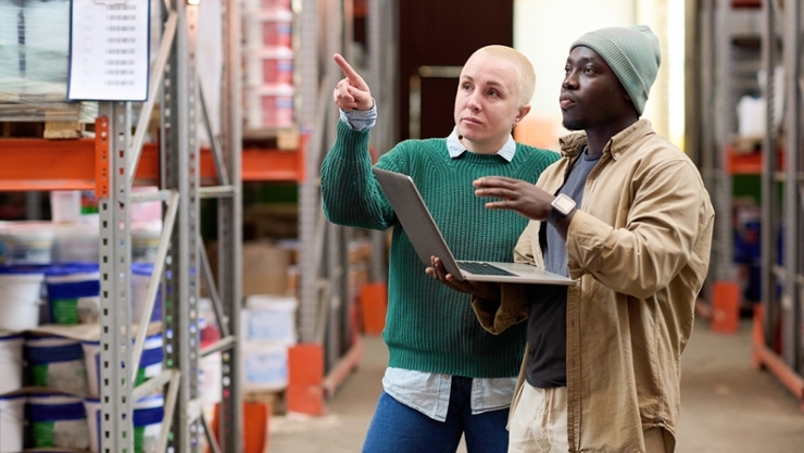 Eine Frau und ein Mann mit einem Laptop führen eine Gefährdungsbeurteilung in einem Warenlager durch
